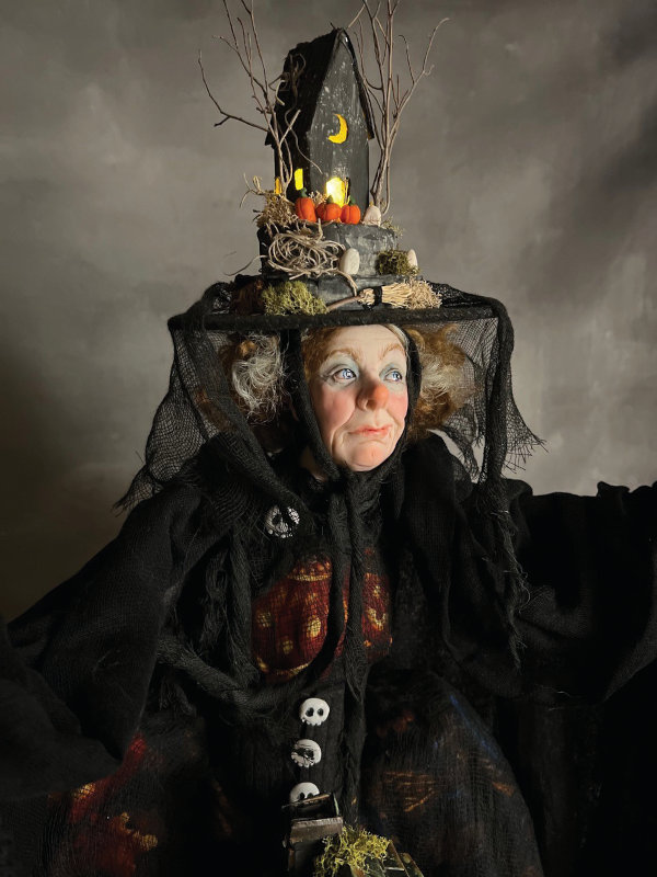 Her witch hat is a miniature haunted house scene that lights up, and she stands over another, larger, haunted house that also lights up.