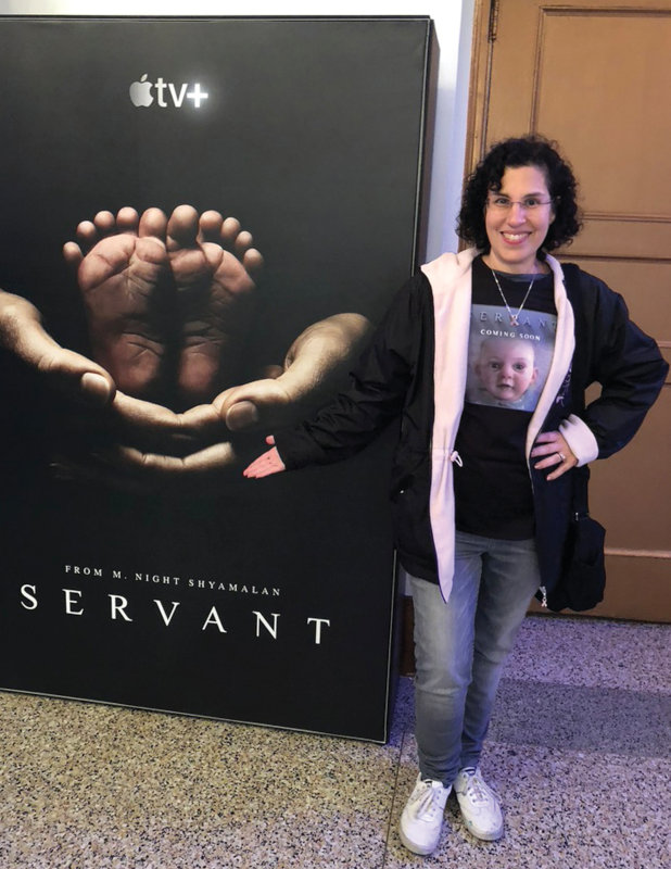 Tuzio-Ross poses next to a promotional poster for the Apple TV+ streaming series Servant, featuring her doll’s lifelike feet.