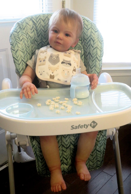 Vito, a silicone baby doll, seated on a high chair with a sippy cup and snacks on the tray in front of him.