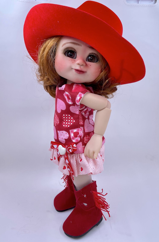 Doll wearing a red dress with heart print along with red cowboy hat and fringed boots.