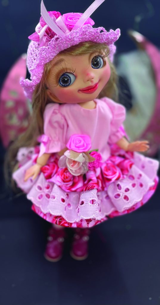 Small BJD wearing a pink dress and matching hat decorated with flowers.