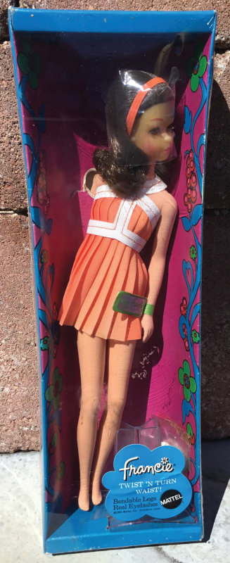 A Mattel Francie Twist 'n Turn Waist doll from 1971, still in its box. The doll has long brunette hair with no bangs and wears a orange dress with white trim.