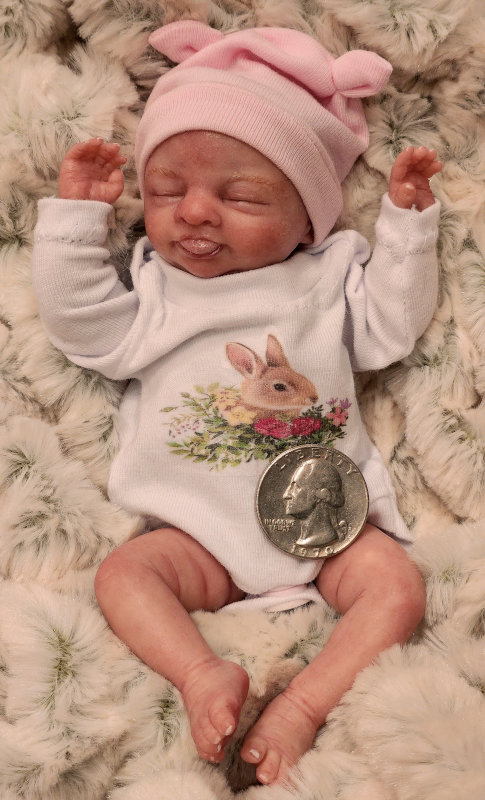 Realistic baby doll posed with a quarter to show the scale - the doll's forearm from wrist to elbow is about the same length as the coin's diameter.