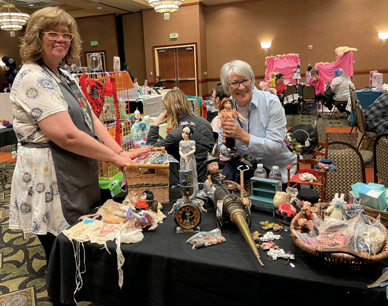 Two women at a convention sales booth. The woman behind the booth holds a doll and is the former owner of the convention.