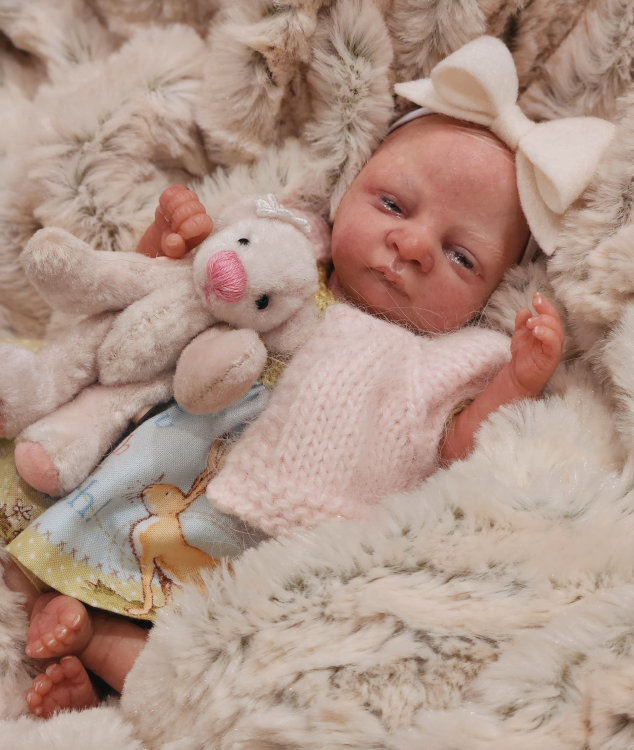 A realistic baby girl doll wearing a sweater over her dress and a bow on her head and holding a stuffed animal.