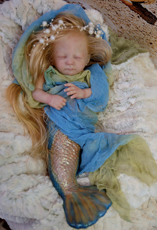 A baby mermaid doll with long blond hair with pearl ornaments, wrapped in gauzy blue and green fabrics.