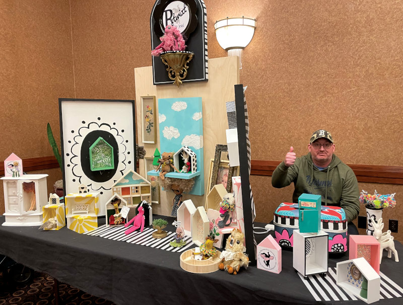 A table displaying room boxes and other doll accessories. A man sitting behind the table is giving a thumbs up for the camera.