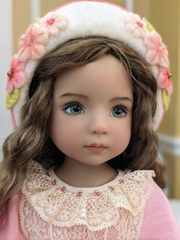 Close-up of a doll from Effner Dolls. She has green eyes and streaky blond hair. She's wearing a pink and white felt hat decorated with felt flowers and a pink dress with cream lace at the neck.