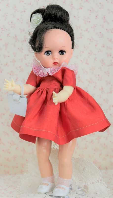Plastic doll with brunette hair in an updo, wearing a red dress with white lace at the neck and white shoes.