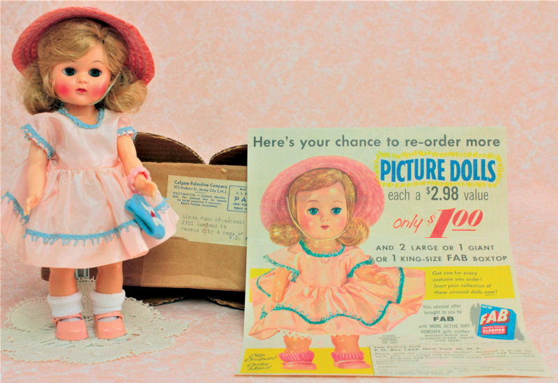 Plastic doll in pink dress with blue trim and pink straw hat standing next to the original box and the ad depicting the doll.