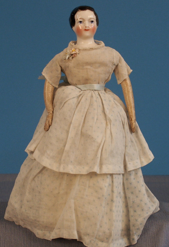 Antique doll with a glazed porcelain head with molded dark brown hair, blue eyes, and rosy cheeks. She wears a long, faded dress with a ribbon at the waist and lace at the neckline. Her arms are pink leather.
