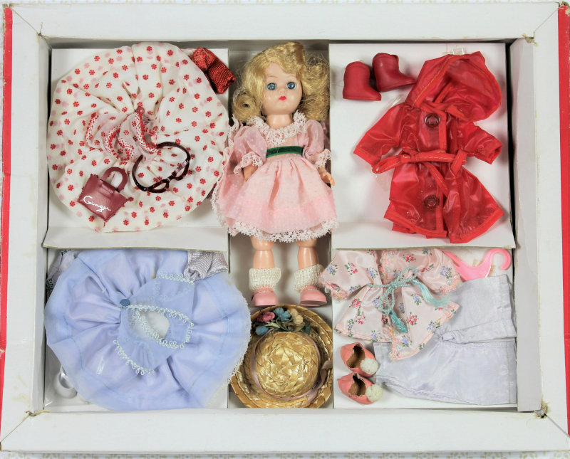 Open box contains a blond plastic doll wearing a pink dress with a straw hat at her feet. Compartments on either side of the doll and hat contain four additional outfits and accessories.
