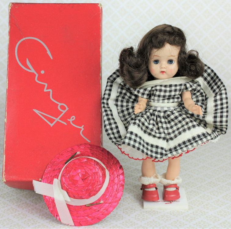 Plastic doll of a long-haired brunette girl wearing a black-and white checked dress and red shoes. A pink straw hat and doll box with "Ginger" logo are to the side.