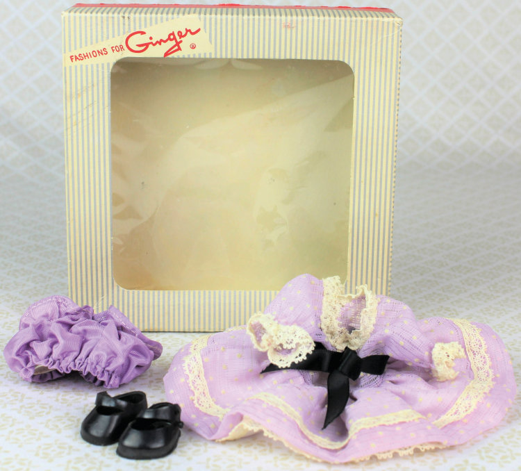 Empty "Fashions for Ginger" box behind a doll dress that's purple with white trim and black ribbon at the waist, purple bloomers, and black shoes.