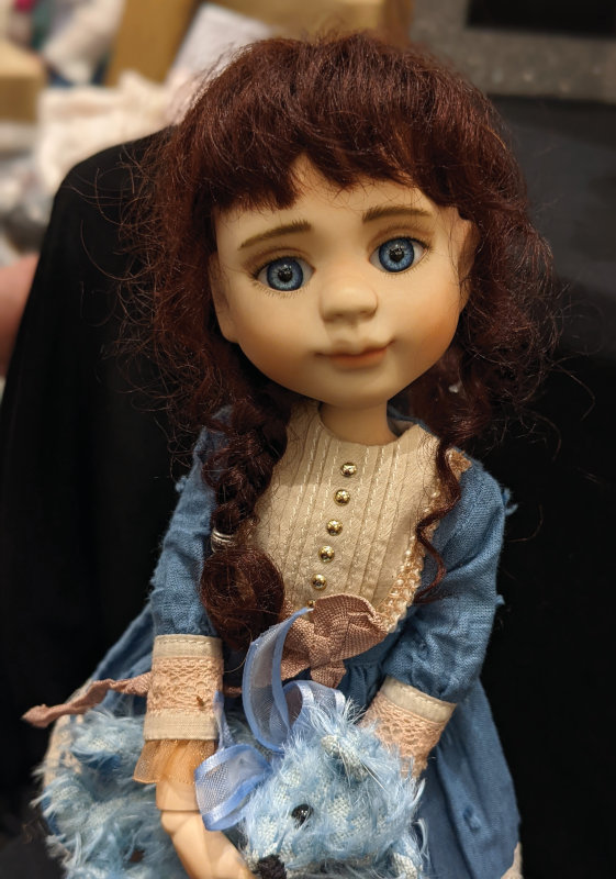 A BJD by Annette Herrman. She has blue eyes and reddish-brown hair in braids and wears a blue dress with cream-colored bodice and trim at the end of her sleeves. She holds a blue teddy bear.