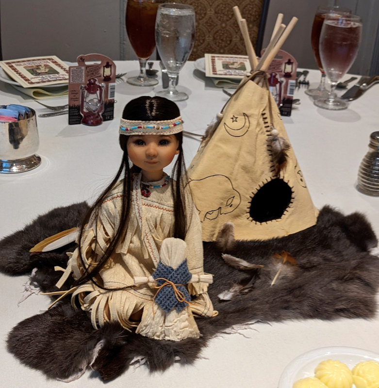 A BJD dressed as a Native American girl posed in front of a tepee in the center of a table.