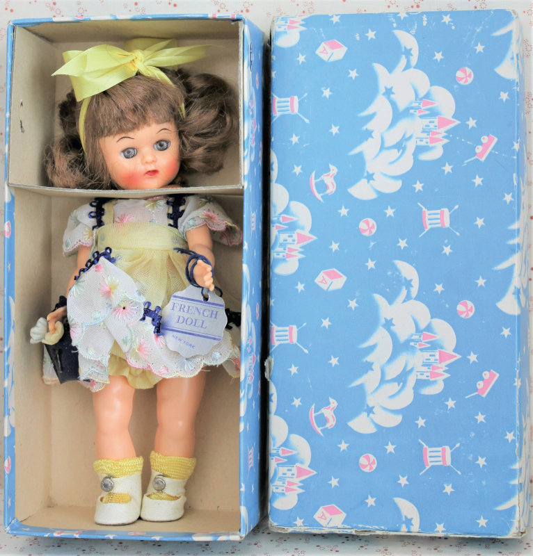 A plastic doll from the 1950s, still in the original box, wearing a dress and hair ribbon with a wrist tag reading "French Doll"