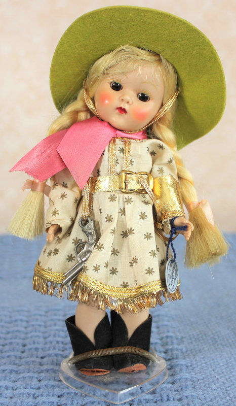 Plastic doll of a child with blond braids wearing a Western-style outfit with green wide-brimmed hat, fringed dress, toy gun, and cowgirl boots.