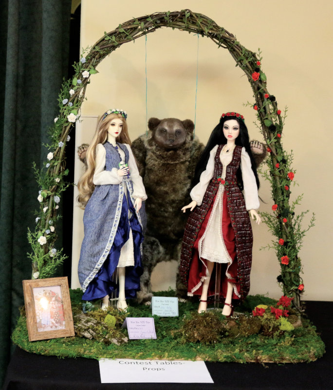 BJDs dressed as Snow White and Rose Red posed on either side of a bear figure, under a floral arch. A sign on the table in front of the scene reads "Contest Tables - Props"