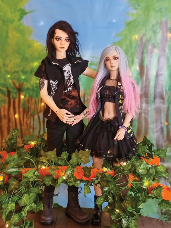 A boy and girl BJD posed holding hands in front of a woodsy backdrop with an ivy-covered fence in front of them.