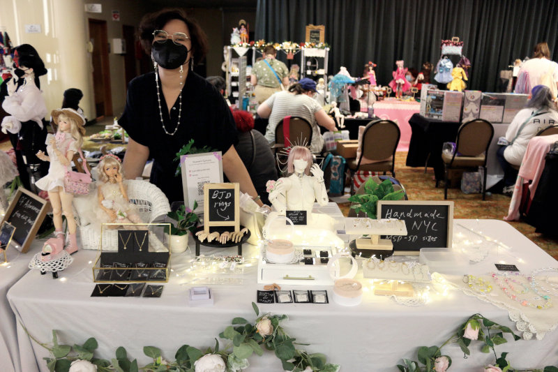 A vendor stands behind her table which dispays several BJDs as well as rings and other doll-sized jewelry.