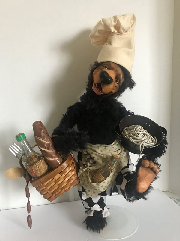A black bear wearing a chef's hat, black-and-white-checkered pants, and an apron carries a pot of noodles and a basket with additional food and serving utensils