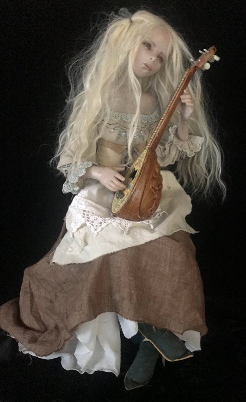 A doll with long, blond hair wears a brown skirt with white apron. She's sitting and holding a mandolin.