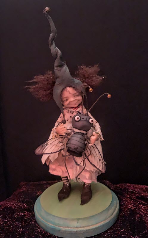 Girl doll with a tall, pointy hat stands on a wood base and holds a fly half her size
