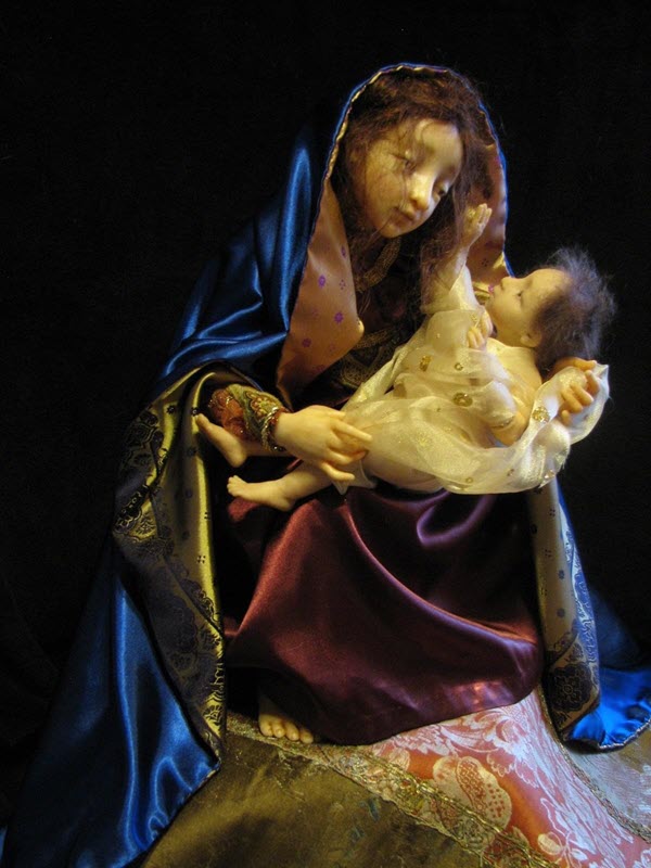Female doll wears a blue cloak and red dress; she holds holds a child doll wearing white, draped clothes.