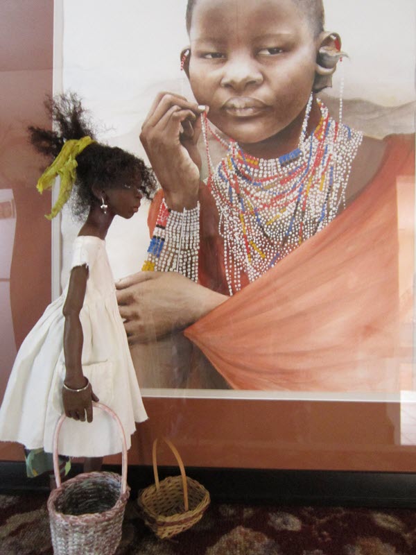 A brown-skinned doll wearing a white, short-sleeved dress and hair ribbon, standing in front of a watercolor painting depicting a person of color wearing traditional beaded jewelry