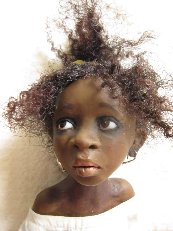 Doll with dark-brown skin tone and textured hair wears a white top. She's looking off to the side.