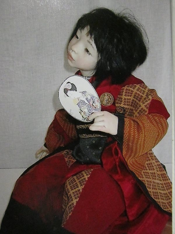 A doll with Asian features wears a red-and-black kimono-style outfit and holds a fan or mirror