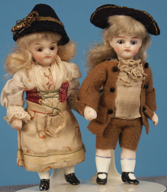 Two blond, porcelain dolls standing next to each other. On the left is a girl, and the right is a boy.
