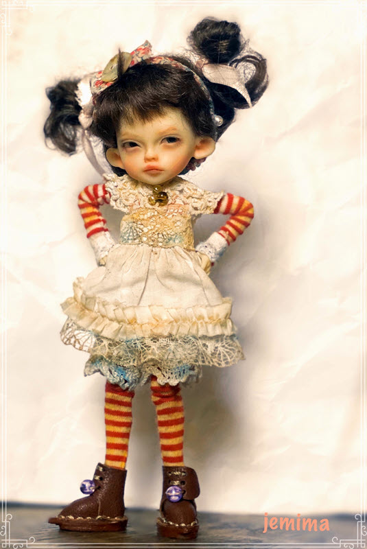 Doll with brown hair in pig tails wears a white lace dress and has red-and-cream-striped clothes covering the arms and legs