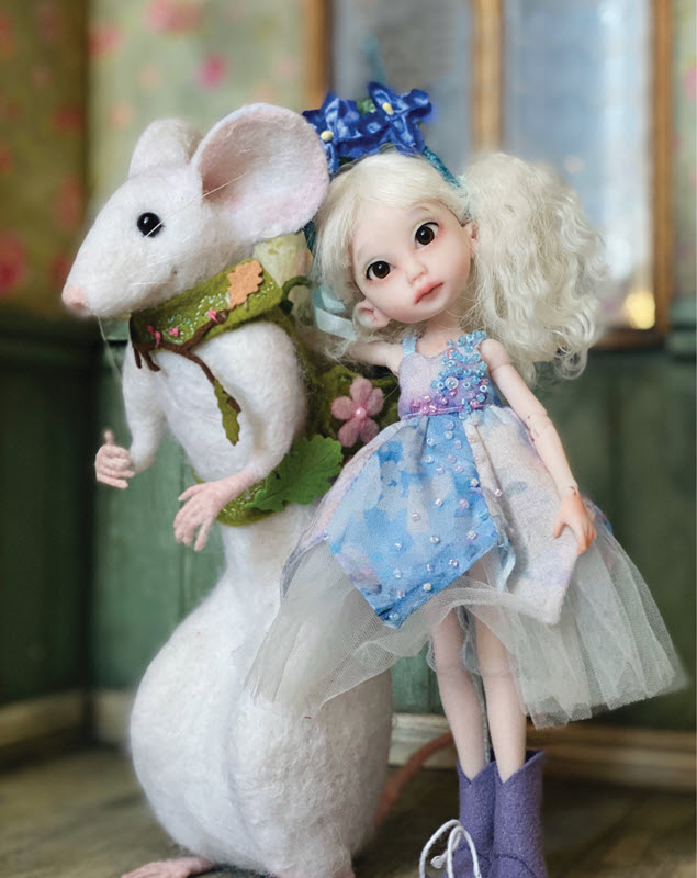 Pale doll with white hair and a bluish dress stands beside a decorative white mouse that is standing on its back legs and wearing a green nature-inspired saddle