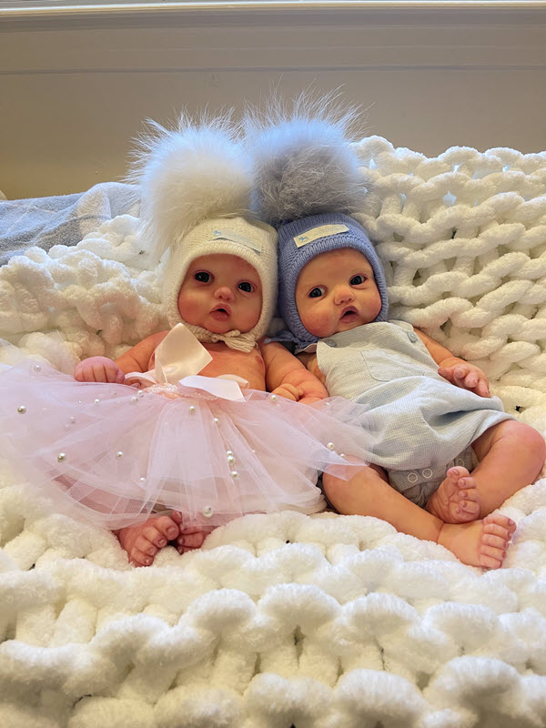 Two baby dolls are posed resting on a white blanket. One wears a pink tutu and the other wears a blue jumper. Both have on knit hats with a large puffball on top.
