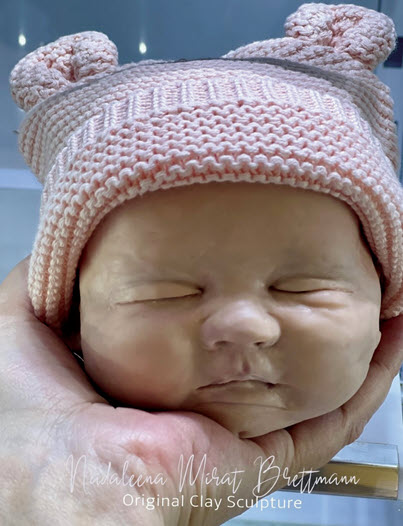 The artist holds the head of a sculpt that is a work in progress and is meant to look like her daughter as an infant. The sculpt has on a knit hat.