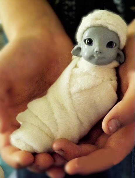 Doll with gray skin and lamb ears is swaddled in white fabric