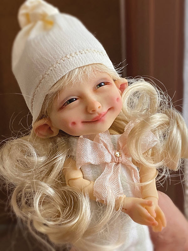 Doll with a girl face is smiling and has on a hat over her long, blond hair