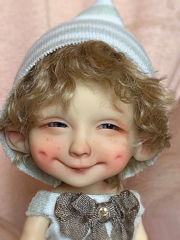Doll with a boy face is smiling and has on a striped hat over his blond hair