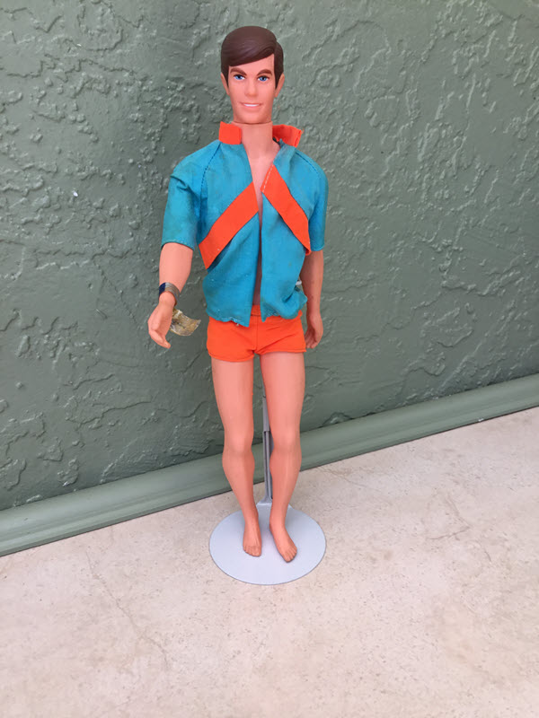 A Ken doll is in a stand, wearing orange shorts and a blue and orange top