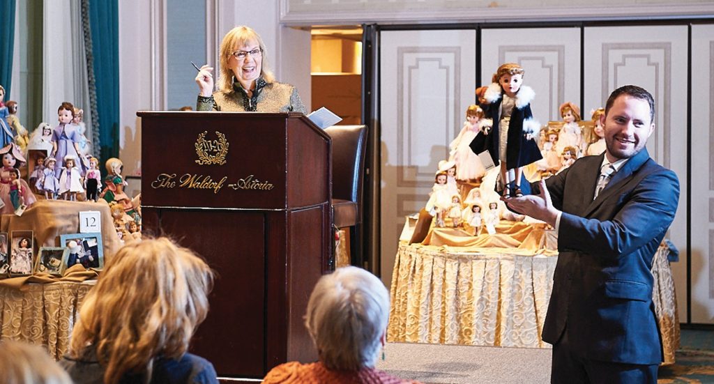 woman at podium facilitating doll auction with a man holding up doll to be bid on