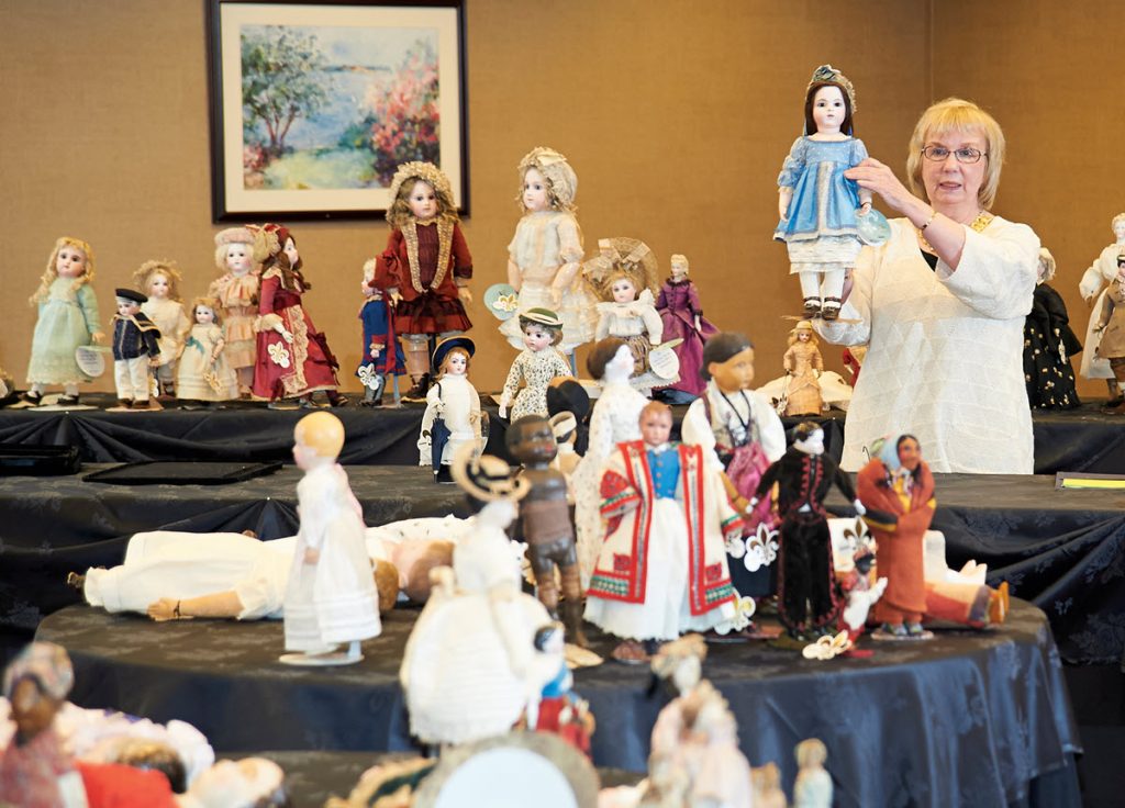 woman surrounded by dolls, holding up doll in blue dress